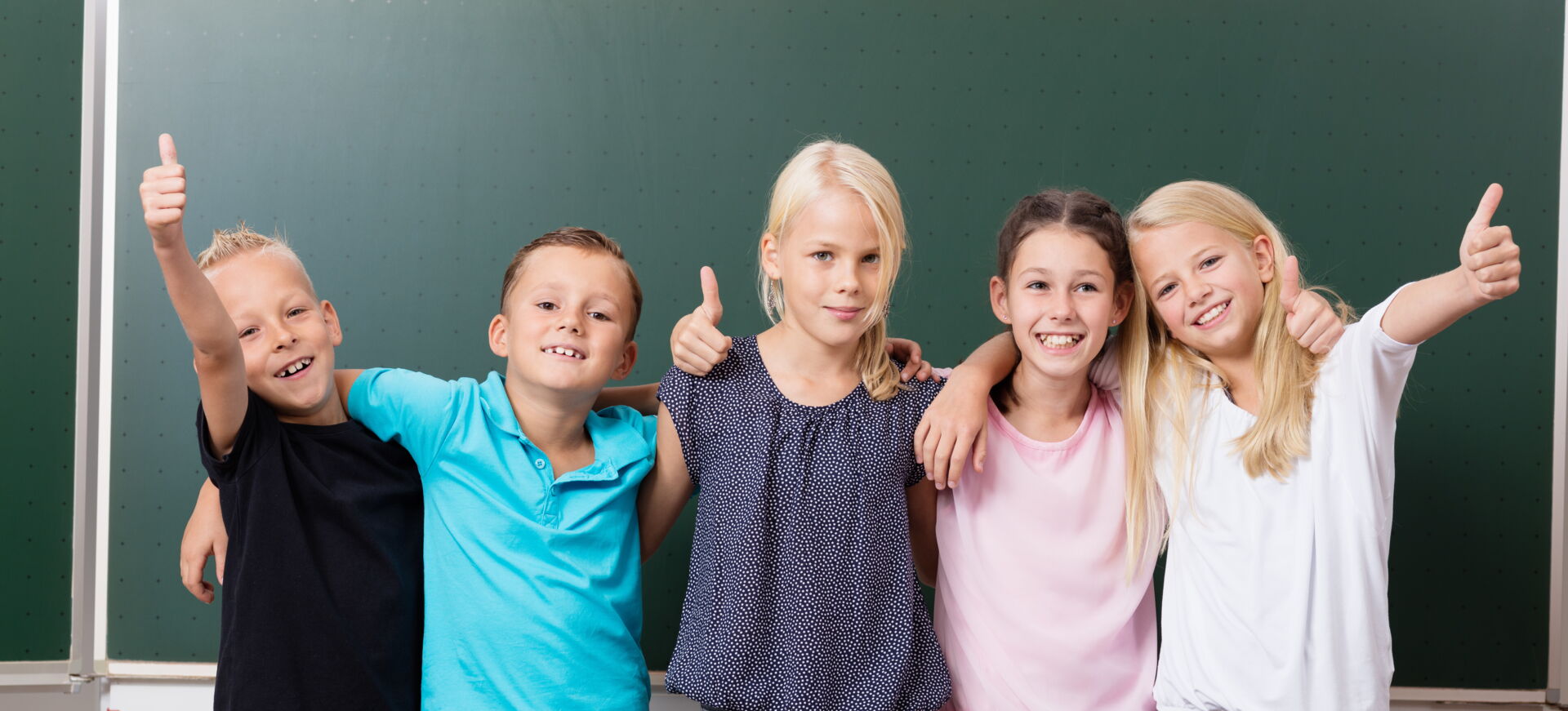 Fünf fröhliche Schulkinder stehen mit erhobenen Daumen hinter einer Schulbank vor einer grünen Tafel. Grundschule „Bjarnat Krawc“ Königswartha.