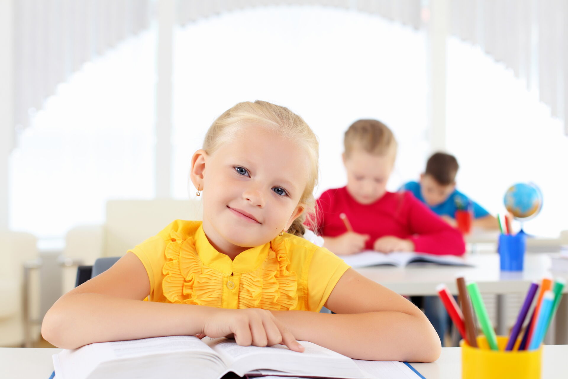 Ein blondes Mädchen in einem gelben Oberteil liest lächelnd in einem Buch in einem hellen Klassenzimmer. Grundschule „Bjarnat Krawc“ Königswartha.