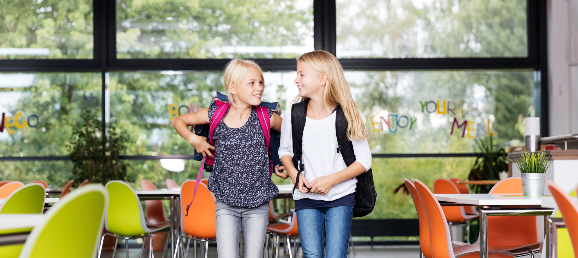 Zwei blonde Mädchen mit Rucksäcken unterhalten sich fröhlich in einem hellen Schulgebäude. Grundschule „Bjarnat Krawc“ Königswartha.