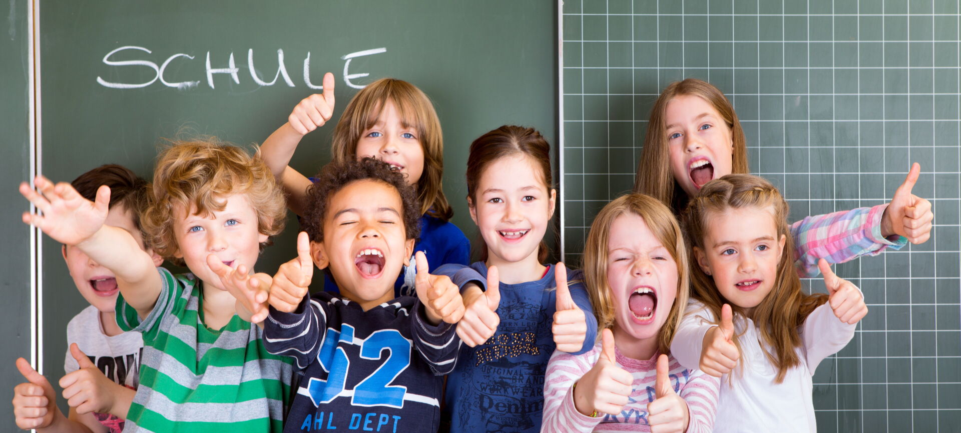 Fröhliche Schulkinder unterschiedlichen Alters und Geschlechts zeigen vor einer grünen Tafel mit der Aufschrift "SCHULE" den Daumen nach oben. Grundschule „Bjarnat Krawc“ Königswartha.