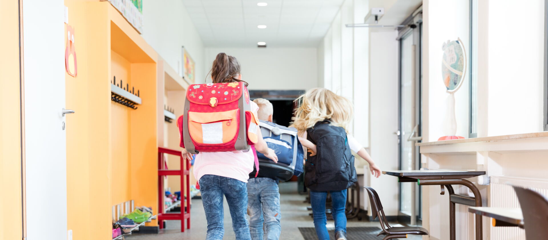 Drei Kinder mit bunten Schulranzen rennen einen hellen Flur in Richtung eines Klassenzimmers. Grundschule „Bjarnat Krawc“ Königswartha.